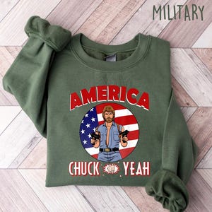 May include: Military green sweatshirt with the word "AMERICA" in red block letters. The design features an illustration of a man with guns, a red and white striped background, and a blue circle with white stars. The text "CHUCK YEAH" is below the illustration.