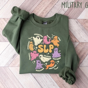 May include: A green sweatshirt with a graphic of ghosts representing different aspects of speech language pathology (SLP). The ghosts are arranged in a circle around the word "SLP" with text labels like "Cognition", "Articulation", "Language", "Hearing", "Voice", "Social", "Stuttering", "Swallowing", and "Motoricities".