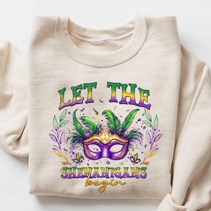 May include: A beige sweatshirt with a green, purple, and gold Mardi Gras mask graphic and the text "Let the Shenanigans begin".