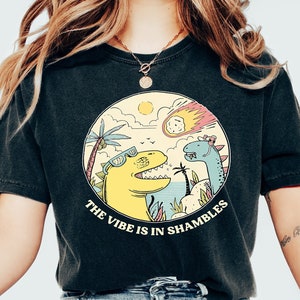 Peut inclure: Un t-shirt noir avec un motif de deux dinosaures dans un d&eacute;cor tropical. Le texte "The vibe is in shambles" est imprim&eacute; sous les dinosaures.