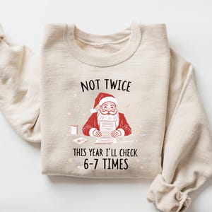 May include: A sand-colored sweatshirt with the text "NOT TWICE THIS YEAR I'LL CHECK 6-7 TIMES" and a graphic of Santa Claus. Santa is wearing a red suit and hat, and is holding a list. The sweatshirt has long sleeves.