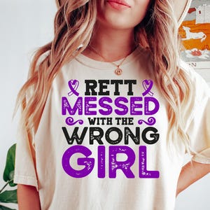 May include: Cream-colored t-shirt with the text "RETT MESSED WITH THE WRONG GIRL" in purple and black. The shirt features a purple ribbon design. The model is wearing dark green shorts and a bracelet.