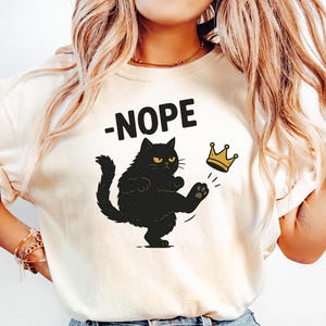 May include: Cream-colored t-shirt with the word "-NOPE" above a black cat kicking a gold crown. The cat has yellow eyes and is in a dynamic pose. The shirt is paired with light blue jeans.