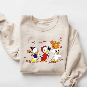May include: A beige sweatshirt featuring a printed design of three cartoon ducks dressed in Halloween costumes. The ducks are wearing a witch hat, a devil costume, and a ghost costume. The design is surrounded by orange and red leaves.