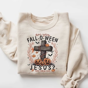 May include: A beige sweatshirt with a graphic print of a wooden cross with pumpkins and bats. The text on the sweatshirt reads "Are you Fall-O-Ween Jesus? Matthew 4:19".