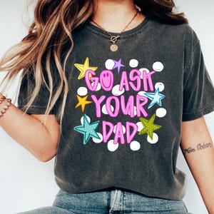 May include: Dark gray t-shirt with the phrase "GO ASK YOUR DAD" in pink and white bubble letters, accented by colorful star graphics. The shirt is paired with blue jeans and white sneakers.