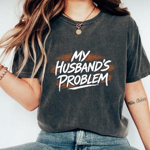 May include: Dark gray t-shirt with the phrase "MY HUSBAND'S PROBLEM" in white lettering over a brown brushstroke design. The shirt is paired with blue jeans and white sneakers, creating a casual, everyday look.