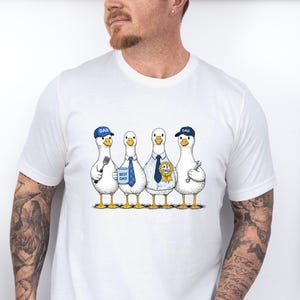May include: White t-shirt featuring a cartoon illustration of four geese. Each goose is dressed in a different outfit, including a baseball cap that says "DAD", ties, and a shirt with a dog. The geese are standing in a row.