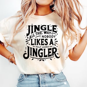 May include: A cream-colored t-shirt with the text "JINGLE ALL THE WAY, NOBODY LIKES A HALF-ASSED JINGLER" in black. The shirt is paired with light blue jeans. The overall theme is a humorous take on the holiday season.