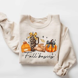 May include: A beige sweatshirt with a graphic design of a pumpkin, boots, a hat, and a leopard print tumbler. The text "Fall basics" is printed below the graphic.