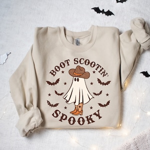 May include: A beige sweatshirt with a graphic of a ghost wearing a cowboy hat and boots. The text "BOOT SCOOTIN' SPOOKY" is printed on the sweatshirt.