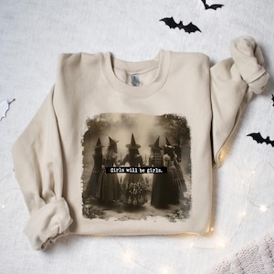 May include: A beige sweatshirt with a vintage-style image of five witches in a circle, with the text "Girls will be girls." printed below the image.