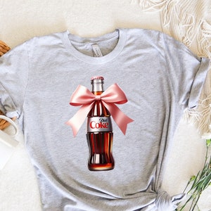 May include: Gray t-shirt with a graphic of a Diet Coke bottle tied with a pink bow.