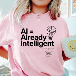 May include: A light pink t-shirt with the text "AI = Already Intelligent" and a brain inside a lightbulb graphic. The shirt also features the text "powered by curiosity" and the SIOHAN PRESS logo. A white mug with a yellow smiley face is held in the hand.