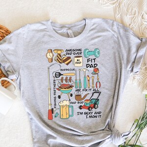 May include: Gray t-shirt with a graphic design of a dad doing various activities, including fishing, barbecuing, mowing the lawn, and fixing things. The text on the shirt reads "Awesome Dad Ever", "#1 Dad", "Fit Dad", "Mr. Fix It All", "Dad Bod", and "I'm Sexy and I Mow It".