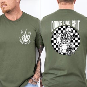 May include: Olive green t-shirt with a white graphic on the front of a skeleton hand with lightning bolts and the text "DOING DAD SHIT". The back of the shirt features a skeleton on a toilet with a phone, surrounded by a black and white checkered circle, and the text "DOING DAD SHIT".