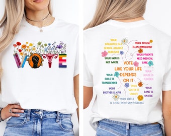 May include: White t-shirt with the word "VOTE" in rainbow colors and a fist and uterus symbol. The back of the shirt has a message about voting and social justice issues.