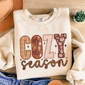 May include: A cream-colored sweatshirt with the text "Cozy season" in brown, pink, and orange letters with fall-themed patterns. The letters are outlined in dark brown.