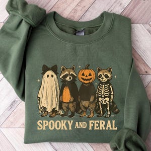 May include: Military green sweatshirt with a Halloween-themed graphic. The design features a ghost, two raccoons, and a pumpkin-headed figure. The text "Spooky and Feral" is printed below the characters. The sweatshirt is folded on a light wood-grain surface.