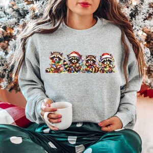 May include: A gray sweatshirt with a Christmas-themed design featuring five raccoons wearing Santa hats and holding Christmas lights. The raccoons are arranged in a line, with the first raccoon on the left and the last raccoon on the right.