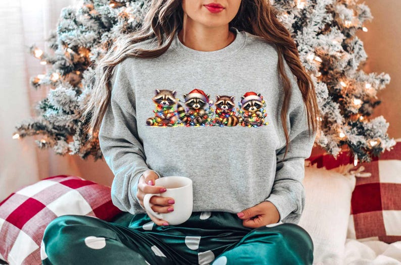 May include: A gray sweatshirt with a Christmas-themed design featuring five raccoons wearing Santa hats and holding Christmas lights. The raccoons are arranged in a line, with the first raccoon on the left and the last raccoon on the right.