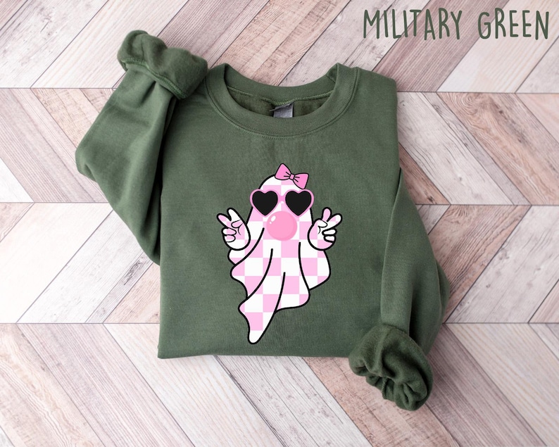 K&ouml;nnte beinhalten: Ein gr&uuml;nes Sweatshirt mit einem rosa-wei&szlig; karierten Geist, der eine Sonnenbrille und eine rosa Schleife tr&auml;gt. Der Text "MILITARY GREEN" befindet sich in der oberen rechten Ecke des Bildes.