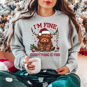 May include: A gray sweatshirt with a Christmas-themed design featuring a Highland cow wearing a Santa hat and Christmas lights. The text "I'm Fine Everything is Fine" is printed on the sweatshirt.