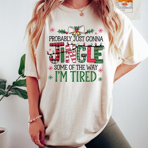 Peut inclure: T-shirt cr&egrave;me avec l'inscription "PROBABLY JUST GONNA JINGLE SOME OF THE WAY I'M TIRED" dans un design festif et color&eacute;. Le mot "JINGLE" est dans un motif &agrave; carreaux avec des guirlandes lumineuses. Le t-shirt est de style d&eacute;contract&eacute; et confortable.