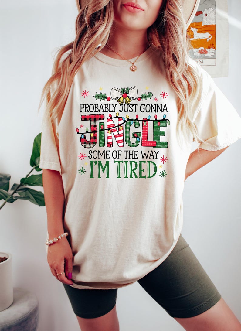 Peut inclure: T-shirt cr&egrave;me avec l'inscription "PROBABLY JUST GONNA JINGLE SOME OF THE WAY I'M TIRED" dans un design festif et color&eacute;. Le mot "JINGLE" est dans un motif &agrave; carreaux avec des guirlandes lumineuses. Le t-shirt est de style d&eacute;contract&eacute; et confortable.