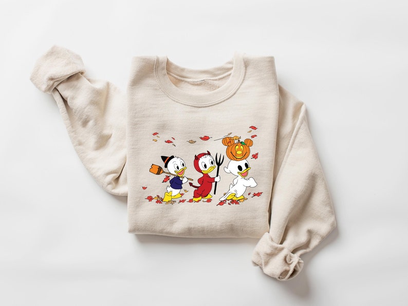 May include: A beige sweatshirt featuring a printed design of three cartoon ducks dressed in Halloween costumes. The ducks are wearing a witch hat, a devil costume, and a ghost costume. The design is surrounded by orange and red leaves.