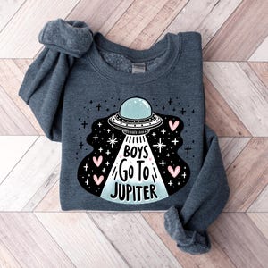 以下が含まれることがあります： 暗いグレーのトレーナーに、UFOのグラフィックと「Boys Go To Jupiter」という文字が白とピンクで書かれています。
