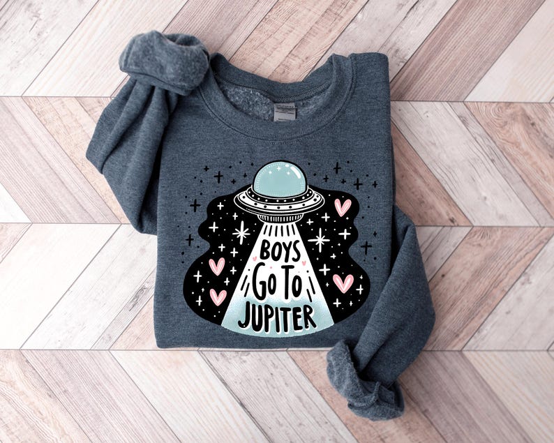 以下が含まれることがあります： 暗いグレーのトレーナーに、UFOのグラフィックと「Boys Go To Jupiter」という文字が白とピンクで書かれています。
