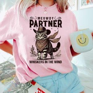 K&ouml;nnte beinhalten: Pinkes T-Shirt mit einer Comic-Katze, die einen Cowboyhut und ein Bandana tr&auml;gt. Der Text auf dem Shirt lautet "MEOWDY PARTNER WHISKERS IN THE WIND".