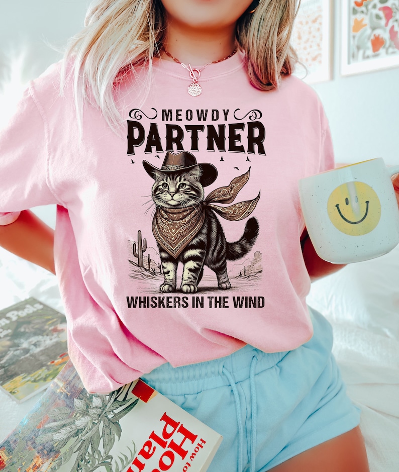 K&ouml;nnte beinhalten: Pinkes T-Shirt mit einer Comic-Katze, die einen Cowboyhut und ein Bandana tr&auml;gt. Der Text auf dem Shirt lautet "MEOWDY PARTNER WHISKERS IN THE WIND".