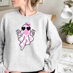 K&ouml;nnte beinhalten: Ein grauer Pullover mit einem rosa-wei&szlig; karierten Geist, der herzf&ouml;rmige Sonnenbrillen und eine rosa Schleife tr&auml;gt. Der Geist macht mit beiden H&auml;nden ein Peace-Zeichen.
