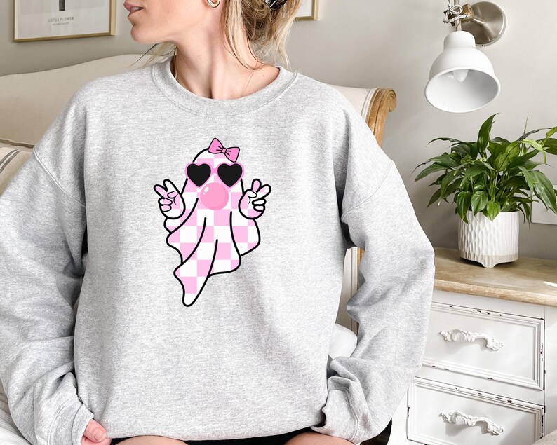 K&ouml;nnte beinhalten: Ein grauer Pullover mit einem rosa-wei&szlig; karierten Geist, der herzf&ouml;rmige Sonnenbrillen und eine rosa Schleife tr&auml;gt. Der Geist macht mit beiden H&auml;nden ein Peace-Zeichen.