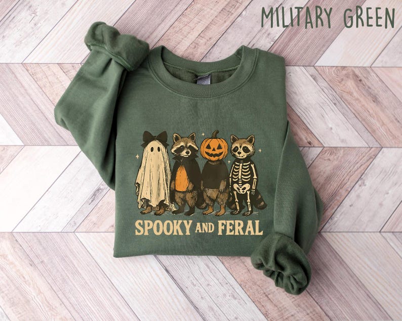 May include: Military green sweatshirt with a Halloween-themed graphic. The design features a ghost, two raccoons, and a pumpkin-headed figure. The text "Spooky and Feral" is printed below the characters. The sweatshirt is folded on a light wood-grain surface.