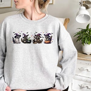 May include: A light grey sweatshirt with a Halloween graphic. The design shows four raccoons in witch hats, with bats and purple details. One raccoon has a cauldron, another sits on a pumpkin. The sweatshirt is a crew neck style, suitable for autumn.
