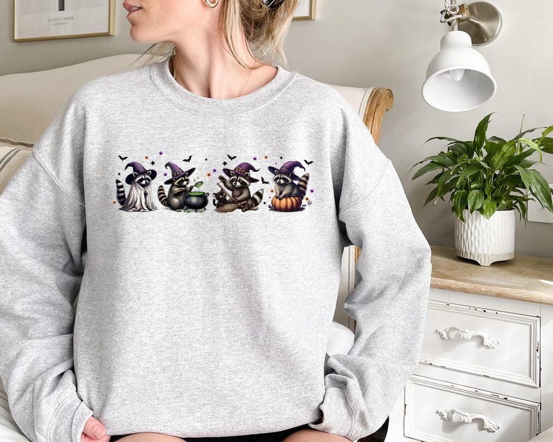 May include: A light grey sweatshirt with a Halloween graphic. The design shows four raccoons in witch hats, with bats and purple details. One raccoon has a cauldron, another sits on a pumpkin. The sweatshirt is a crew neck style, suitable for autumn.