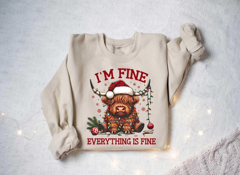 May include: A beige sweatshirt with a Christmas-themed design featuring a Highland cow wearing a Santa hat and wrapped in Christmas lights. The text "I'm Fine Everything is Fine" is printed on the sweatshirt.