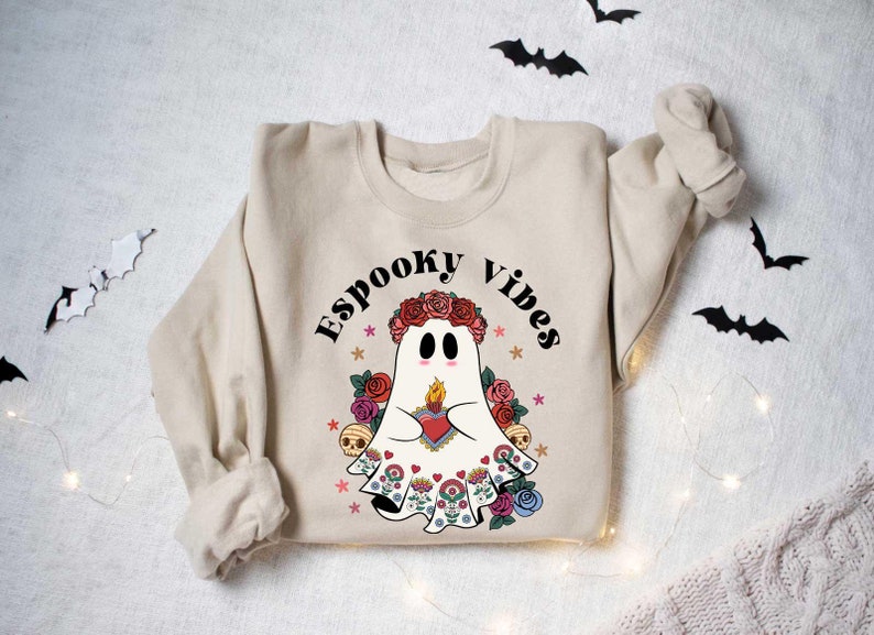 May include: A beige sweatshirt with a white ghost wearing a flower crown and holding a heart with a flame. The text "Spooky Vibes" is printed above the ghost.