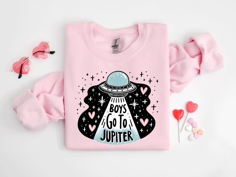 以下が含まれることがあります： ピンクのトレーナーに、UFOと「Boys Go To Jupiter」の文字、ピンクのハートの黒と白のグラフィックが描かれています。