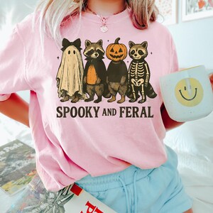 May include: Pink Halloween-themed sweatshirt with the text "SPOOKY AND FERAL." The graphic features four cartoon raccoons dressed as a ghost, vampire, jack-o'-lantern, and skeleton. The person in the image is holding a white mug with a yellow smiley face.