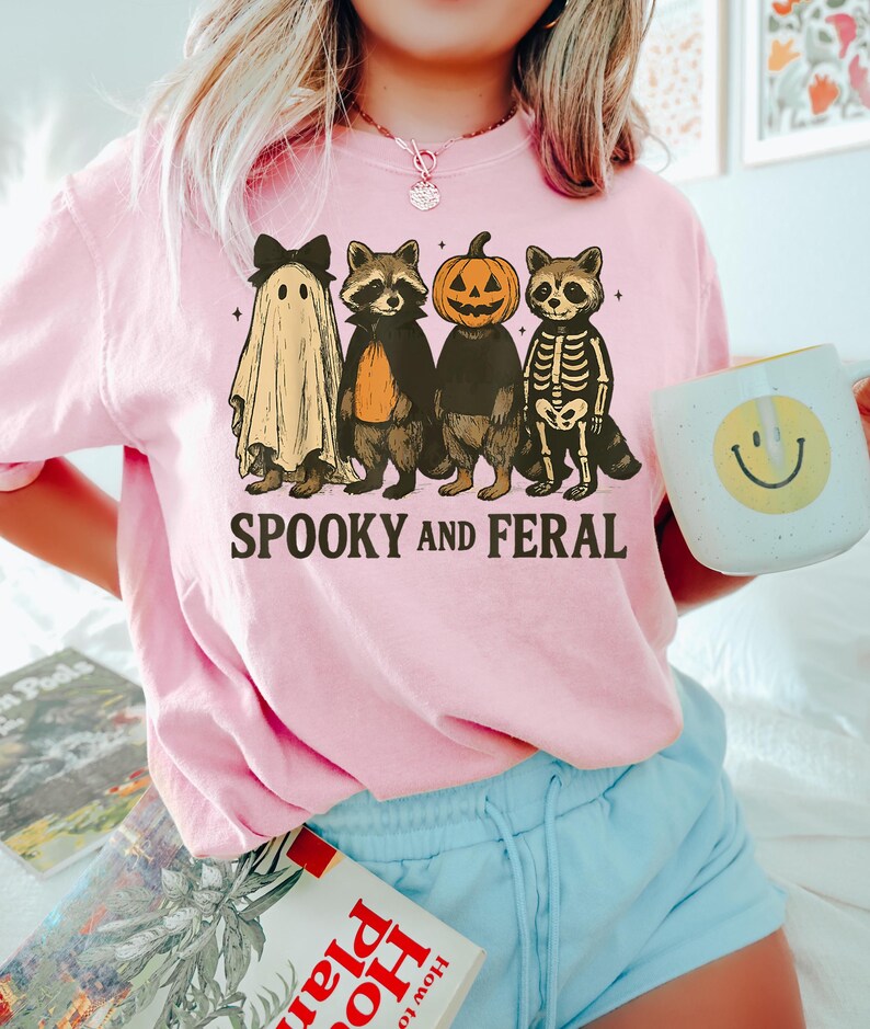 May include: Pink Halloween-themed sweatshirt with the text "SPOOKY AND FERAL." The graphic features four cartoon raccoons dressed as a ghost, vampire, jack-o'-lantern, and skeleton. The person in the image is holding a white mug with a yellow smiley face.