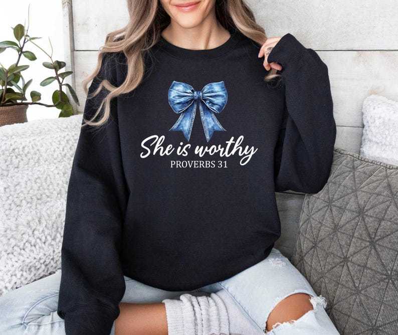 May include: A navy blue sweatshirt featuring a watercolor-style blue bow graphic. The text "She is worthy" and "PROVERBS 31" are printed in white script below the bow. The sweatshirt is a casual garment.