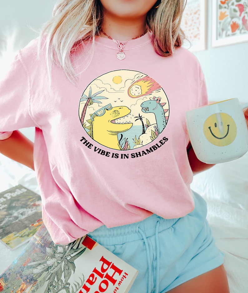 Peut inclure: T-shirt rose avec un motif de deux dinosaures et une com&egrave;te. Le texte sur le t-shirt est "The vibe is in shambles."