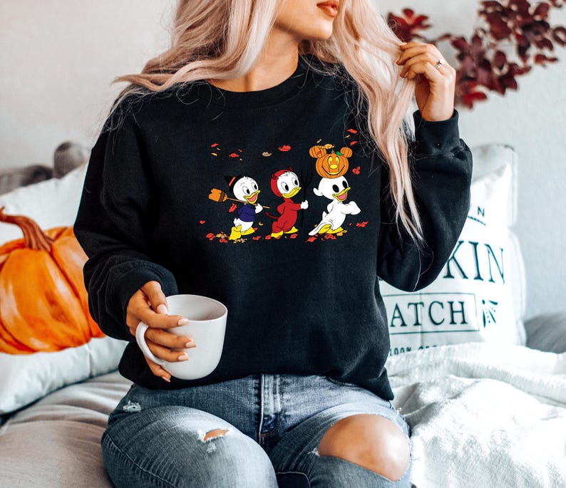 May include: Black sweatshirt featuring a graphic print of three cartoon ducks dressed in Halloween costumes. The ducks are holding a broom, a pumpkin, and a ghost. The sweatshirt has a crew neck and long sleeves.