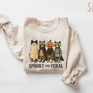 May include: A sand-colored sweatshirt featuring a Halloween-themed graphic. The design includes a ghost, two raccoons, and a pumpkin-headed figure. The text "Spooky and Feral" is printed below the characters. The sweatshirt has long sleeves.