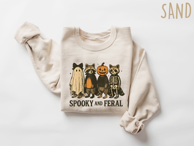 May include: A sand-colored sweatshirt featuring a Halloween-themed graphic. The design includes a ghost, two raccoons, and a pumpkin-headed figure. The text "Spooky and Feral" is printed below the characters. The sweatshirt has long sleeves.