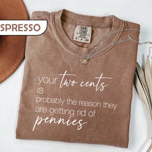 May include: An espresso-colored short-sleeve t-shirt with white text that reads: your two cents IS probably the reason they are getting rid of pennies. The shirt features a crew neckline and a Comfort Colors brand label inside the collar.
